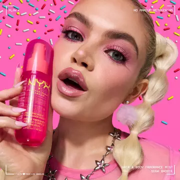 Lifestyle shot of model with dramatic pink eye makeup holding NYX Professional Makeup Suga Baddie Hair & Body Fragrance Mist pink bottle.