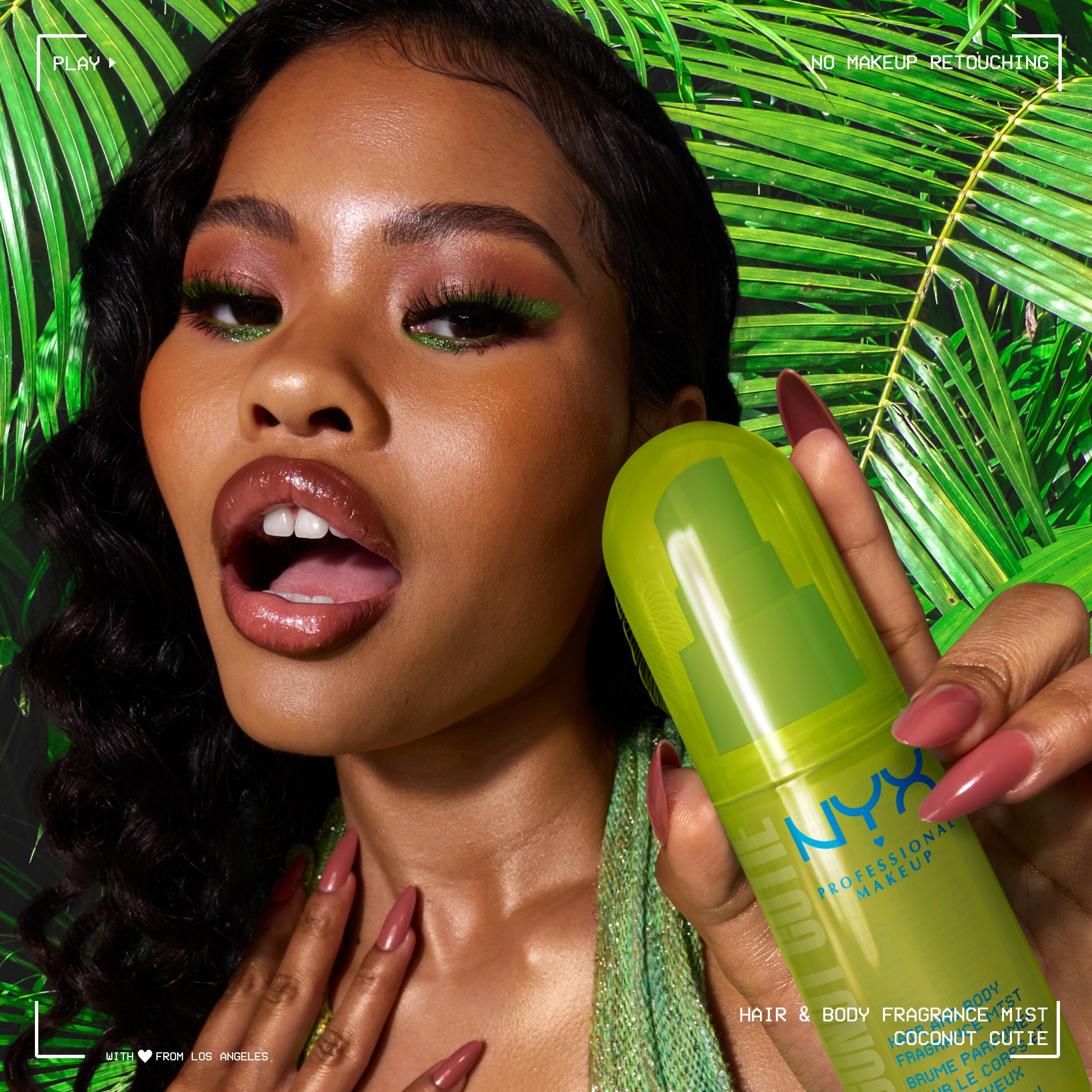 Lifestyle shot of model holding lime green Coconut Cutie Hair & Body Mist with palm leaves in the background.
