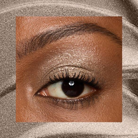 Close-up of a deep-skinned eye wearing 07 Granite Grind, a taupe metallic cream eyeshadow from NYX Professional Makeup Lid Lingerie, on a matching taupe shimmer backdrop.