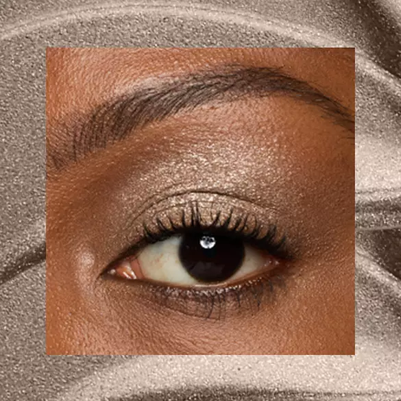 Close-up of a deep-skinned eye wearing 07 Granite Grind, a taupe metallic cream eyeshadow from NYX Professional Makeup Lid Lingerie, on a matching taupe shimmer backdrop.
