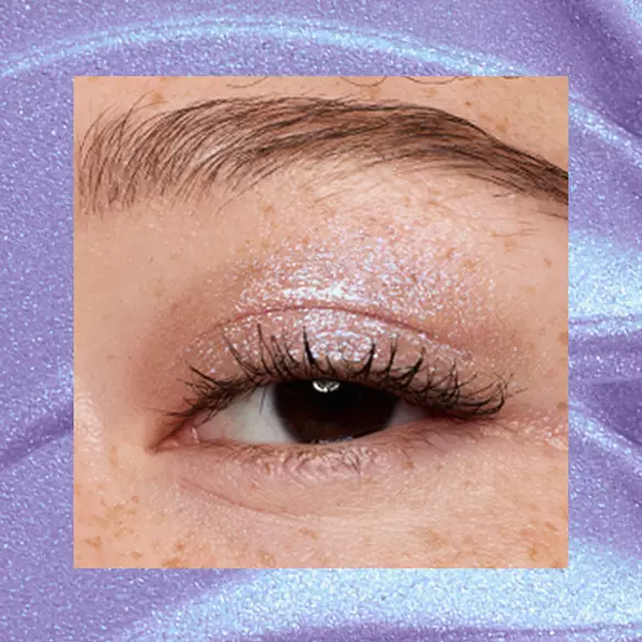 Close-up of a freckled fair-skinned eye wearing 10 Lacy Lilac, a lavender duochrome metallic cream eyeshadow from NYX Professional Makeup Lid Lingerie, on a matching lilac shimmer backdrop.