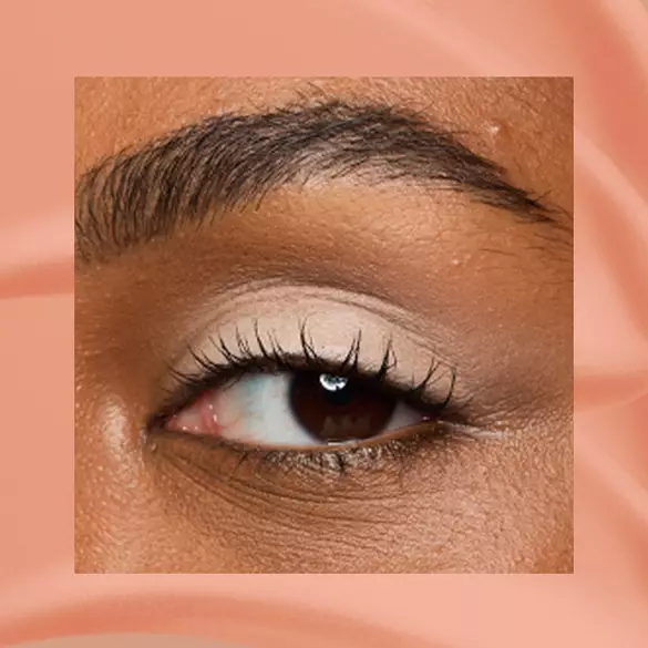 Close-up of a deep-skinned eye wearing 02 In-Nude-Endo, a peachy nude matte cream eyeshadow from NYX Professional Makeup Lid Lingerie, on a matching peach matte backdrop.