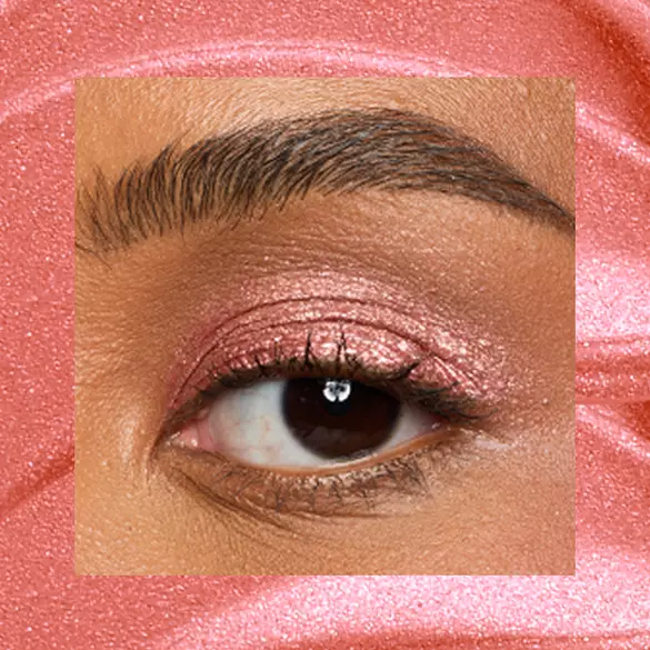 Close-up of a medium-deep-skinned eye wearing 09 Cheeky Coral, a coral pink metallic cream eyeshadow from NYX Professional Makeup Lid Lingerie, on a matching coral shimmer backdrop.